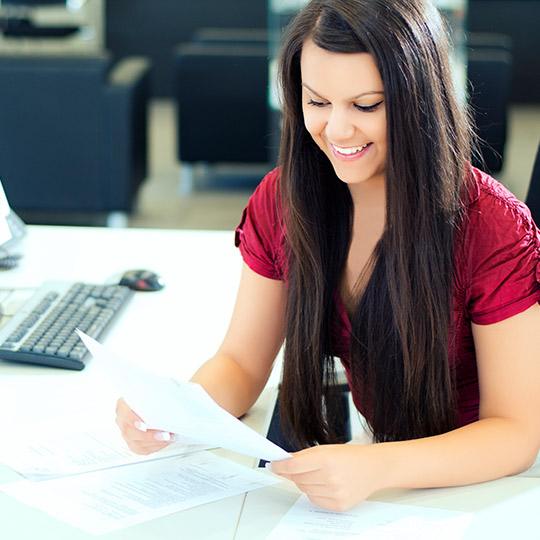 Junge Frau mit langen braunen Haaren liest lächelnd Unterlagen an einem modernen Büroschreibtisch. Junge Frau mit langen braunen Haaren liest lächelnd Unterlagen an einem modernen Büroschreibtisch.